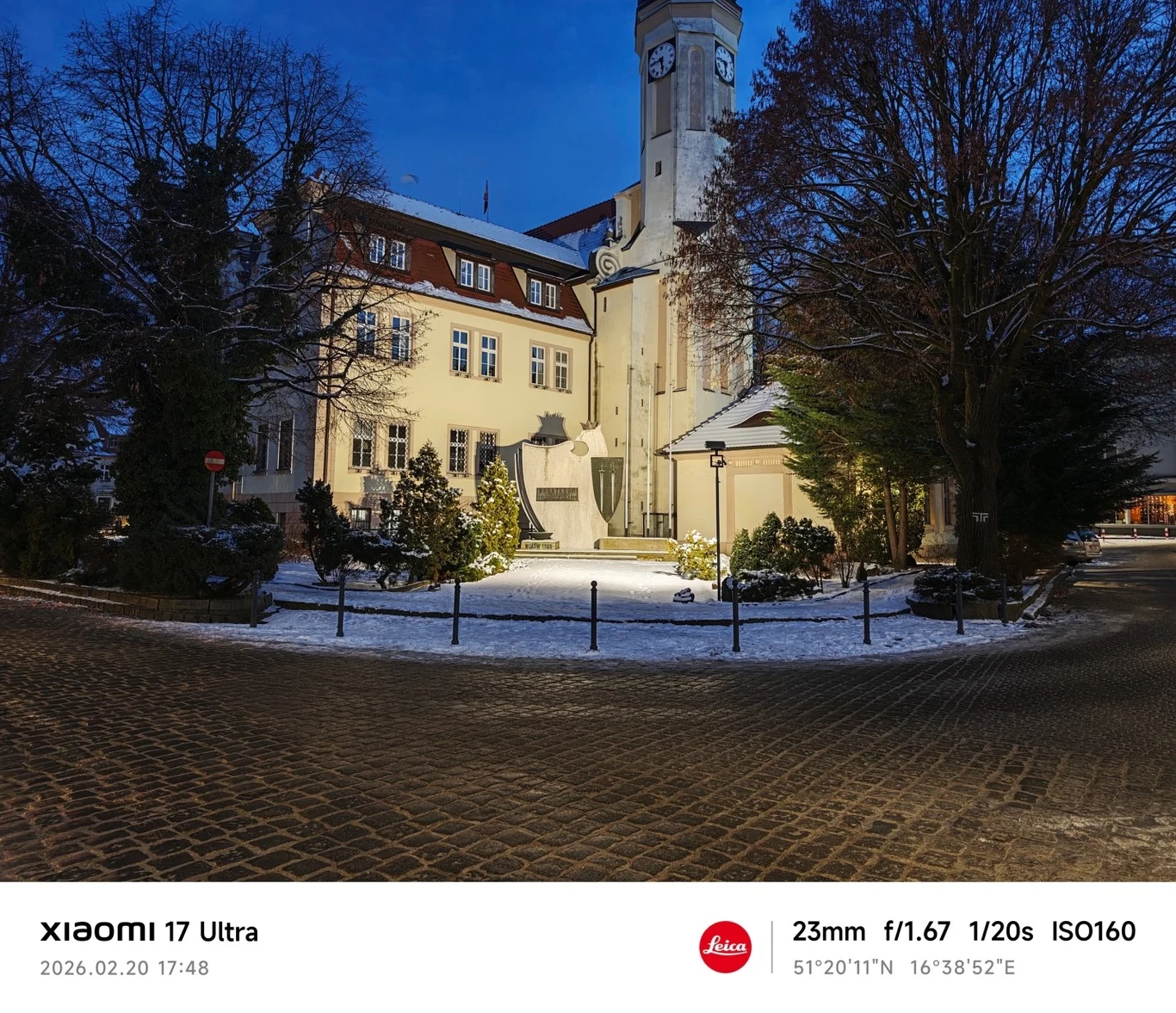 Zabytkowy budynek z wieżą zegarową oświetlony lampami w zimowy wieczór, wokół wybrukowana ulica i przykryte śniegiem chodniki, po bokach drzewa bez liści.