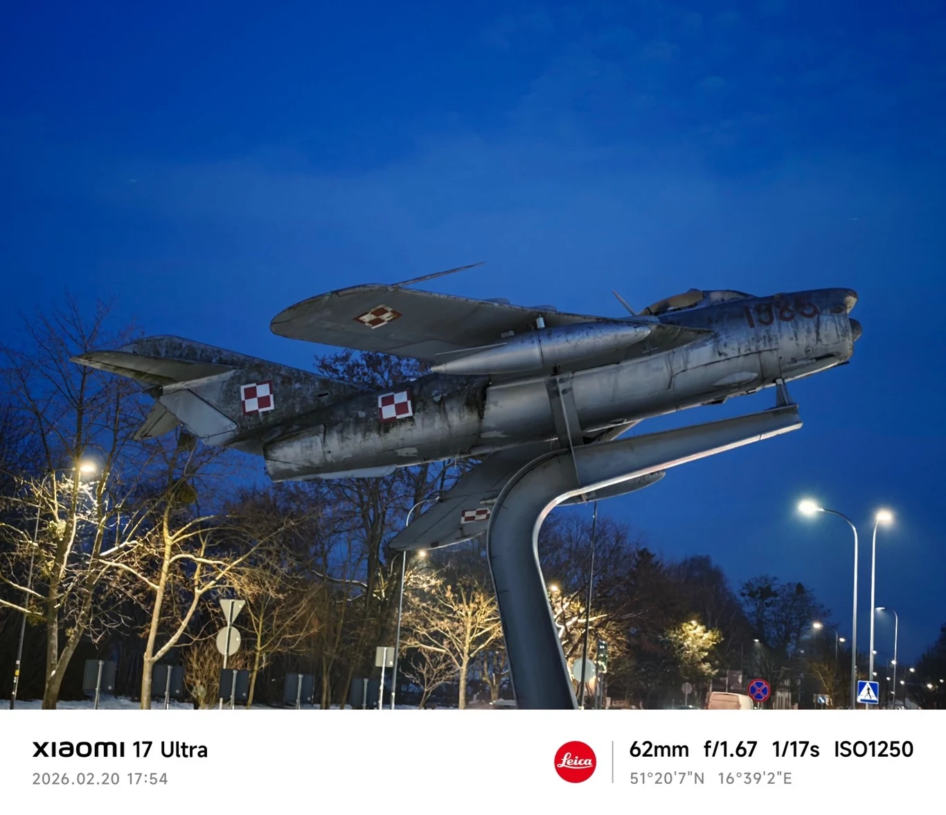Pomnik odrzutowego myśliwca wojskowego z polskimi oznaczeniami lotniczymi umieszczony na metalowej podporze, oświetlony wieczorową porą, w tle drzewa oraz uliczne latarnie.