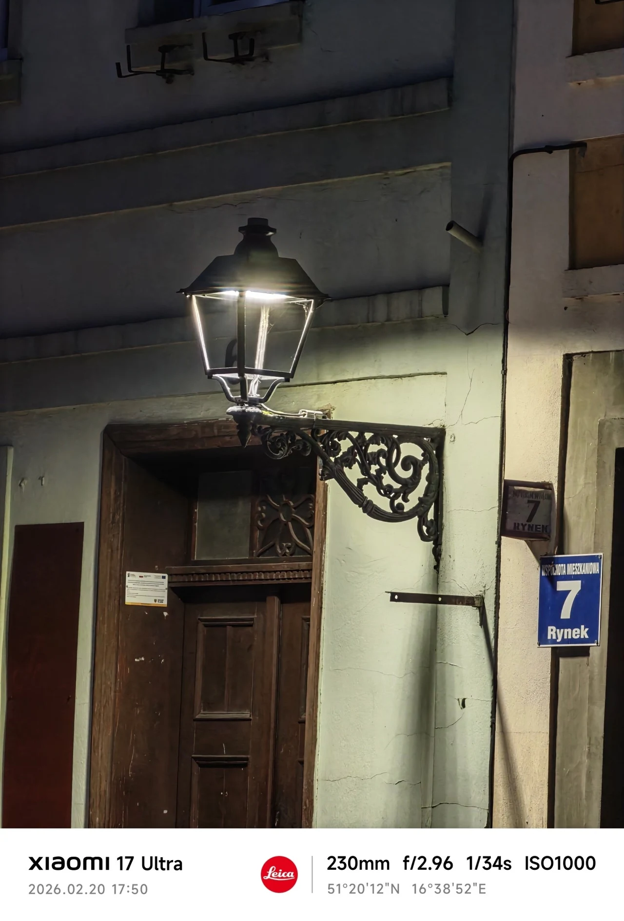 Historyczna uliczna latarnia żeliwna oświetlająca wejście do budynku z drewnianymi drzwiami, obok niebieska tabliczka z numerem 7 i nazwą ulicy Rynek na ścianie.