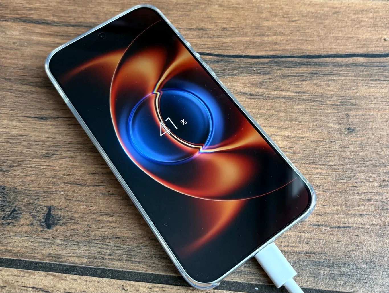 Smartfon z wyświetlaczem ukazującym animację ładowania baterii, położony na drewnianej powierzchni i podłączony do ładowarki przewodowej.