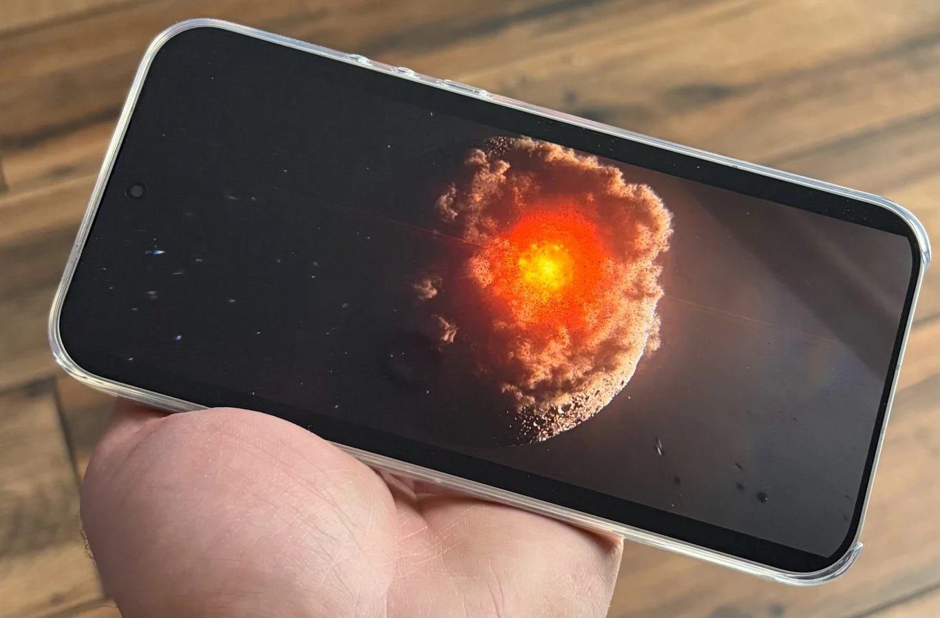 Dłoń trzymająca smartfon, na ekranie którego widoczna jest realistyczna grafika przedstawiająca kosmiczną katastrofę z kulą ognia i eksplozją na tle ciemnej przestrzeni.