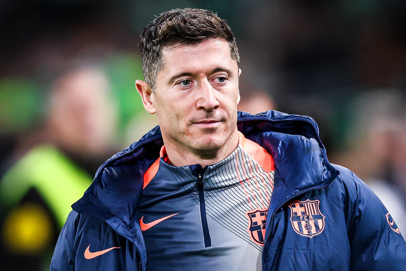 Piłkarz w kurtce sportowej z herbem klubu FC Barcelona, patrzący lekko w bok, w tle rozmyte postacie i zielone kolory trybun.