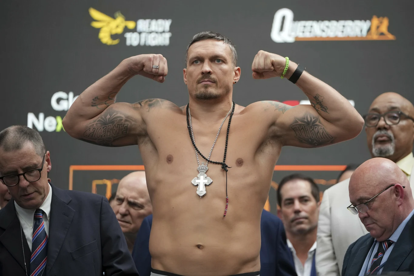 Ołeksandr Usyk, 18.07.2025 r. Mężczyzna z odsłoniętą klatką piersiową, prezentujący umięśnione ramiona w geście zwycięstwa, na szyi duży krzyż, w otoczeniu kilku osób w garniturach podczas oficjalnego wydarzenia sportowego.