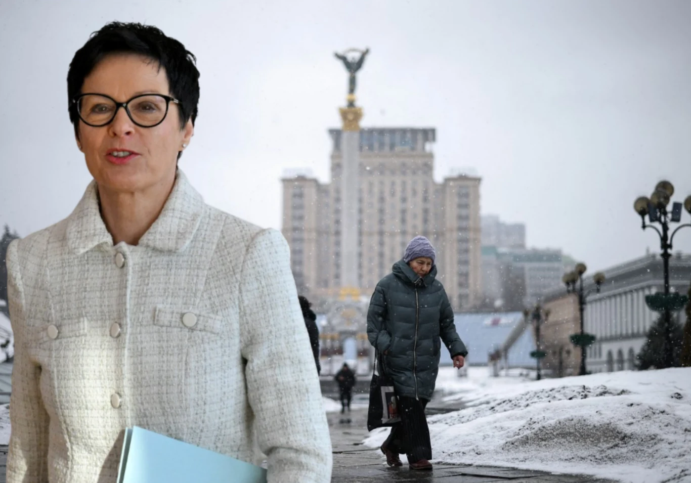 Kobieta w okularach i jasnej marynarce na pierwszym planie, na tle zimowego miasta z widocznym monumentalnym budynkiem oraz pomnikiem anioła, w oddali osoba w ciemnej kurtce spacerująca po zaśnieżonym chodniku.