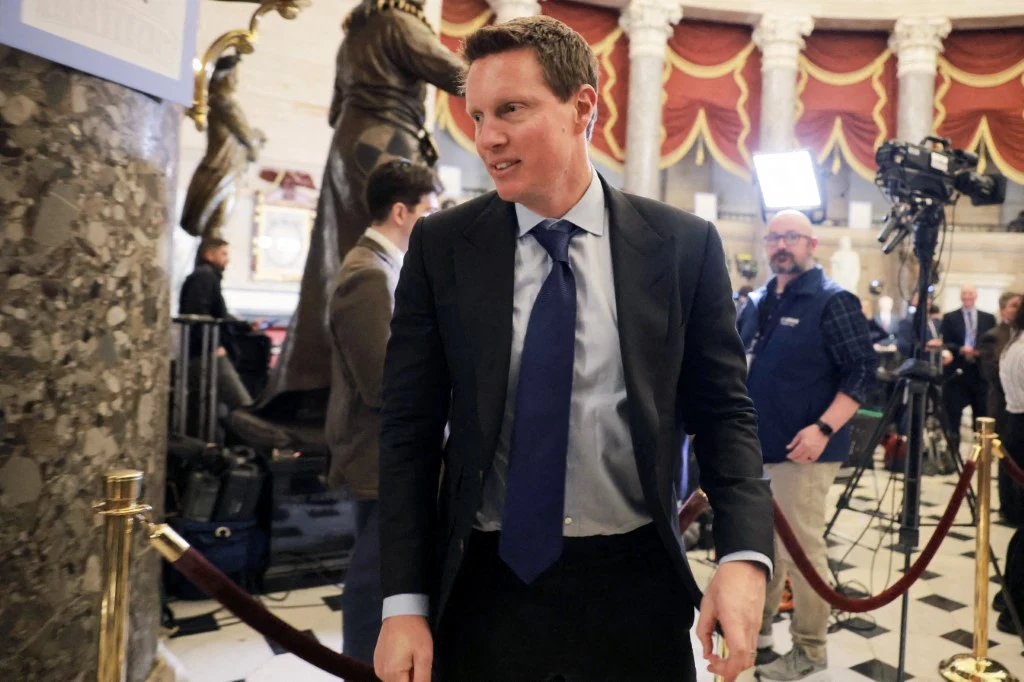 David Ellison Mężczyzna w garniturze i krawacie znajduje się w eleganckim wnętrzu z marmurowymi kolumnami, czerwonymi kotarami i rzeźbami, w otoczeniu ekip dziennikarskich oraz kamer.