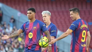 Trzech piłkarzy w koszulkach FC Barcelony znajduje się na murawie stadionu, jeden z nich trzyma piłkę, pozostali dwaj patrzą w jego kierunku, stadion wypełniony jest kibicami.