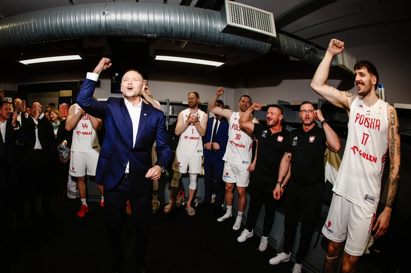 Feta po zwycięstwie "KoszKadry" Koszykarze reprezentacji Polski oraz członkowie sztabu szkoleniowego wiwatują i świętują w szatni, wszyscy wznoszą ręce z okrzykami radości. Część osób ubrana jest w stroje sportowe z nazwą POLSKA, pozostali w garniturach lub czarnych koszulkach.