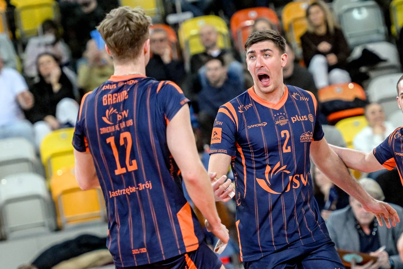 Siatkarze JSW Jastrzębskiego Węgla: Anton Brehme i Łukasz Kaczmarek Dwóch siatkarzy drużyny Jastrzębski Węgiel w granatowo-pomarańczowych strojach podczas emocjonującego momentu meczu na hali sportowej, wokół widoczni kibice na trybunach.