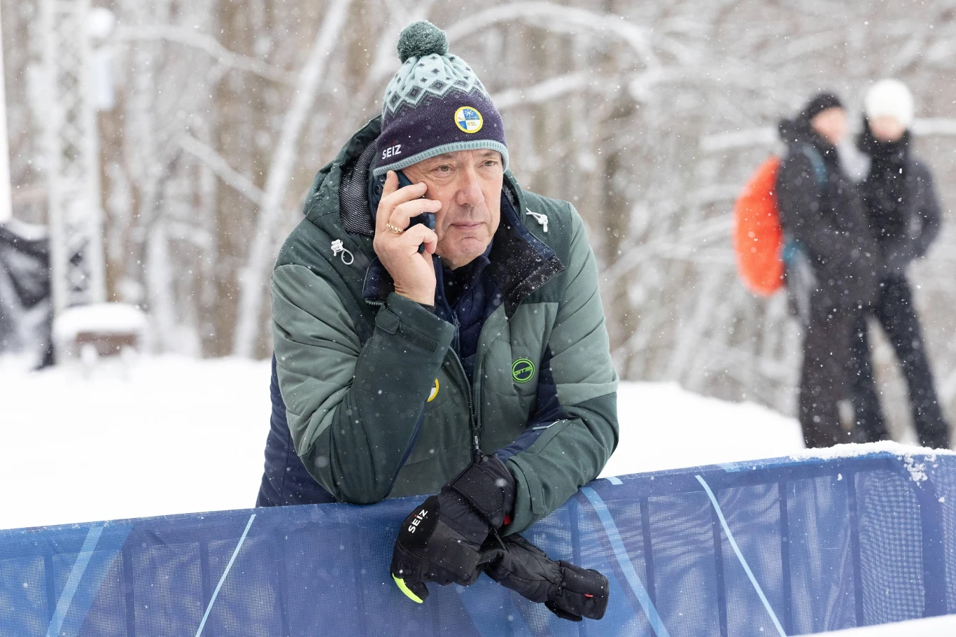 Mężczyzna w zimowej kurtce i czapce rozmawia przez telefon komórkowy, opierając się o barierkę na tle zaśnieżonego lasu, w tle dwie osoby rozmawiające ze sobą.