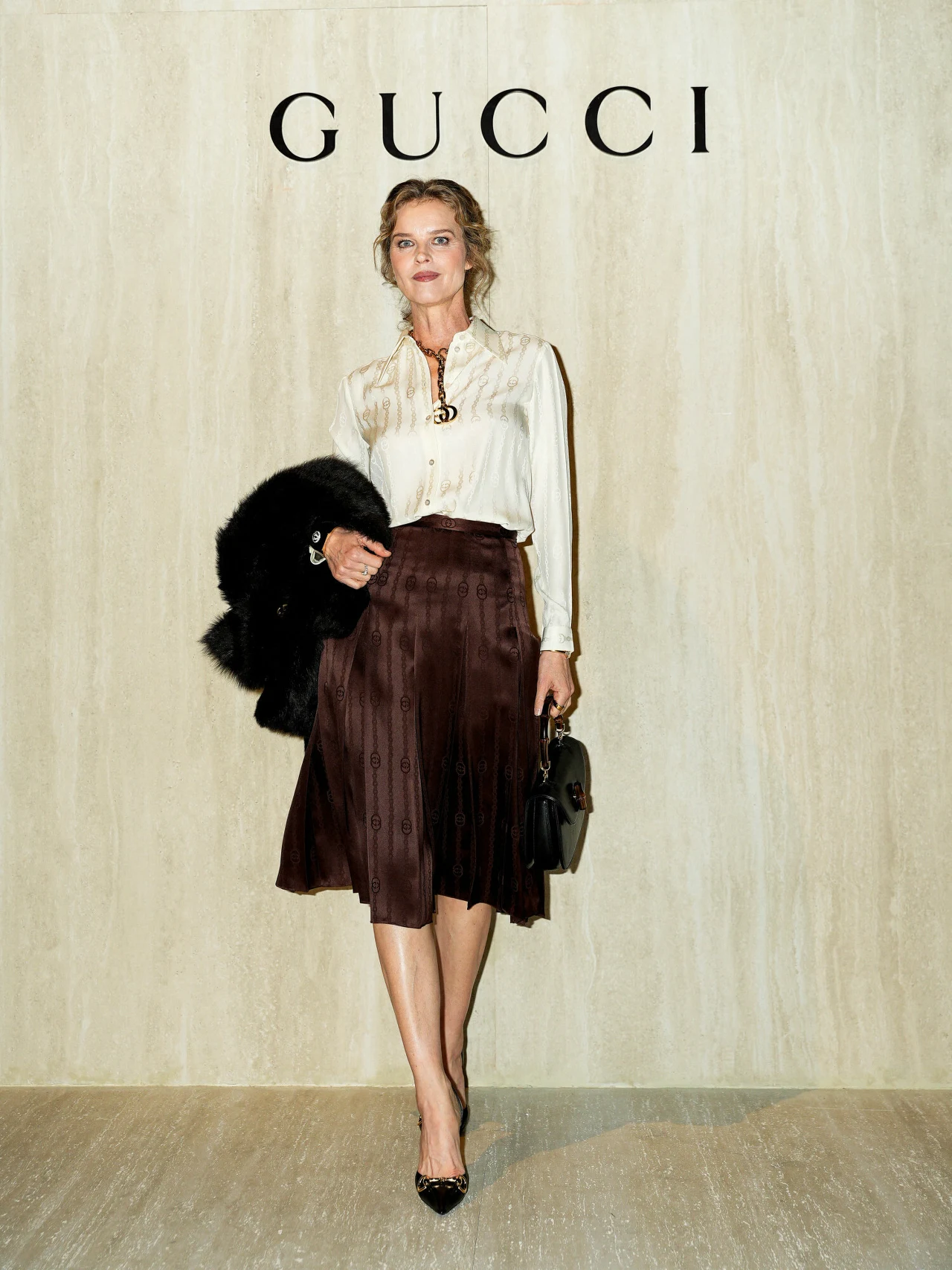 Kobieta ubrana elegancko w kremową bluzkę i bordową spódnicę stoi na tle ściany z dużym napisem Gucci. W jednej ręce trzyma czarną torebkę, w drugiej futrzaną narzutkę, a całość stylizacji dopełniają klasyczne szpilki. Kobieta patrzy wprost przed siebi...