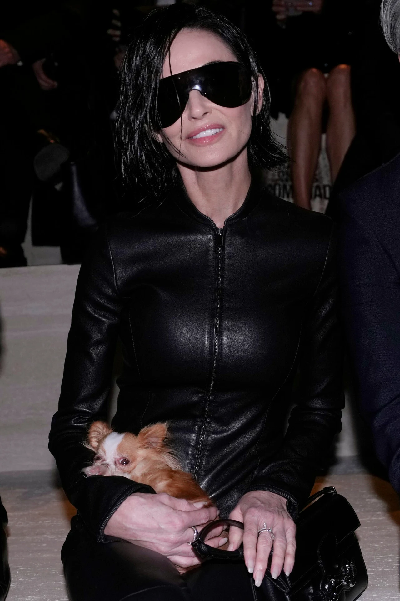 Kobieta w ciemnych okularach przeciwsłonecznych i skórzanej kurtce trzymająca małego psa na kolanach, z delikatnym uśmiechem, siedzi pośród osób na wydarzeniu.