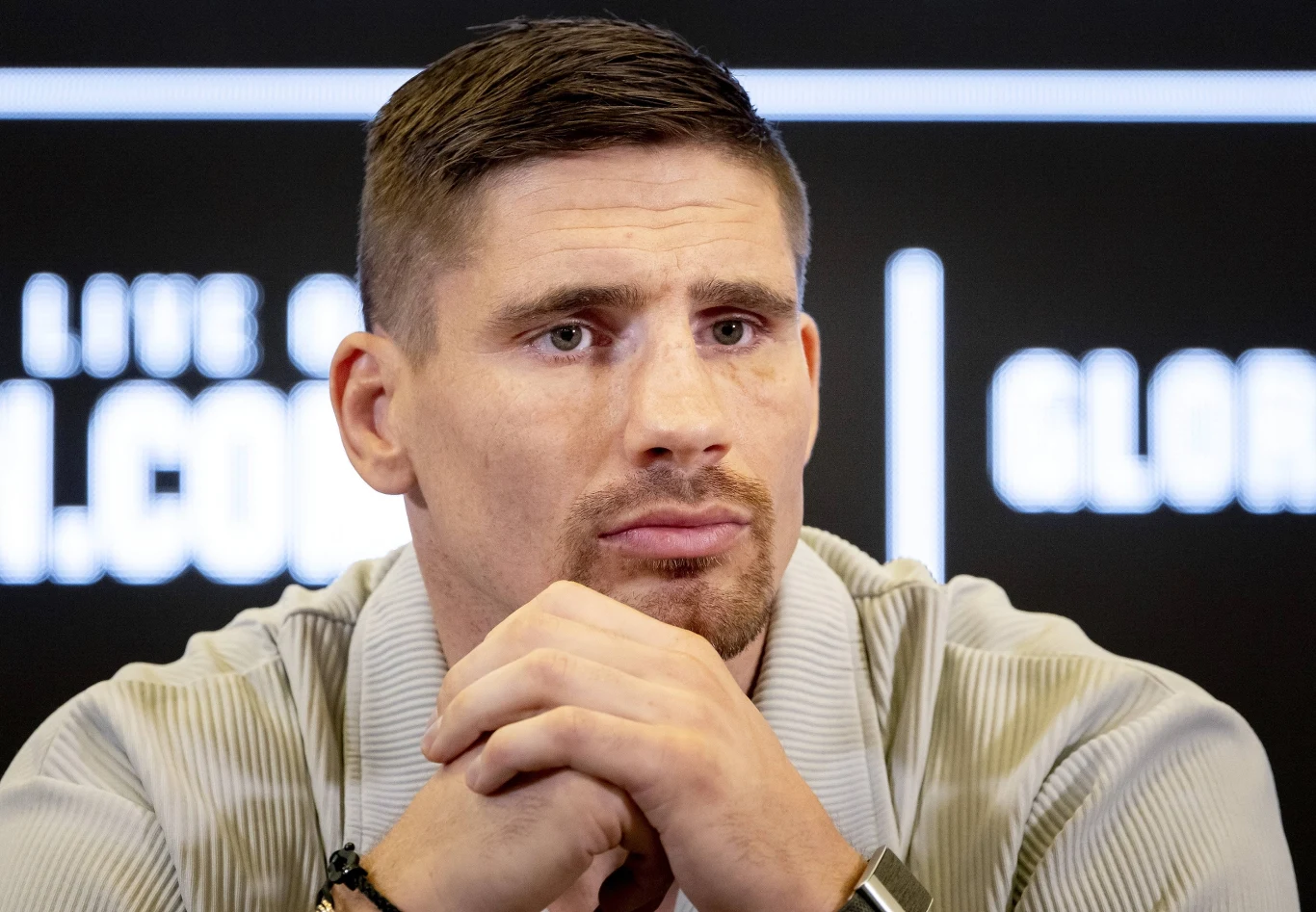 Mężczyzna o poważnym wyrazie twarzy siedzi ze splecionymi dłońmi, ubrany w jasną koszulę, w tle widoczne niewyraźne napisy na ciemnym ekranie.