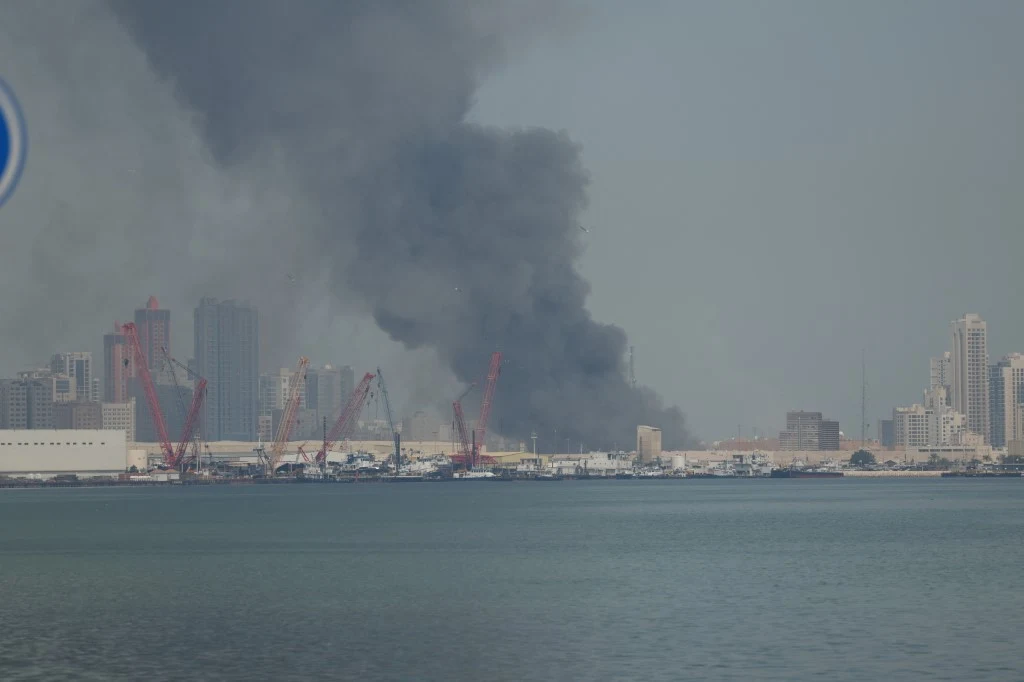 Iran zaatakował amerykańską bazę w Bahrajnie Gęsty, ciemny dym unoszący się nad portem ze strefą przemysłową i licznymi żurawiami, w tle wyłaniają się wysokie budynki miejskiej metropolii.