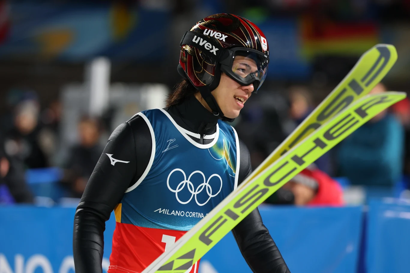 Zawodnik w stroju narciarskim z kaskiem i goglami, trzymający narty marki Fischer. Na stroju widnieje logo olimpijskie oraz napis Milano Cortina, nawiązujący do olimpiady zimowej.