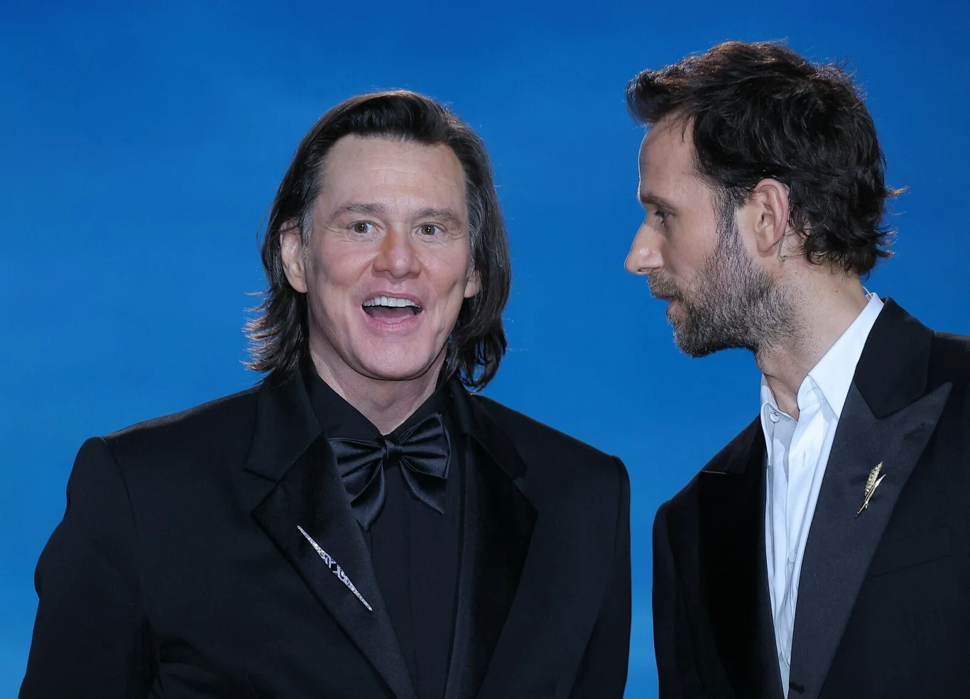 Jim Carrey na Cezarach 2026 Dwóch mężczyzn w czarnych garniturach na tle niebieskiego tła, jeden z nich uśmiechnięty, drugi pochylony w jego stronę jakby szeptał.
