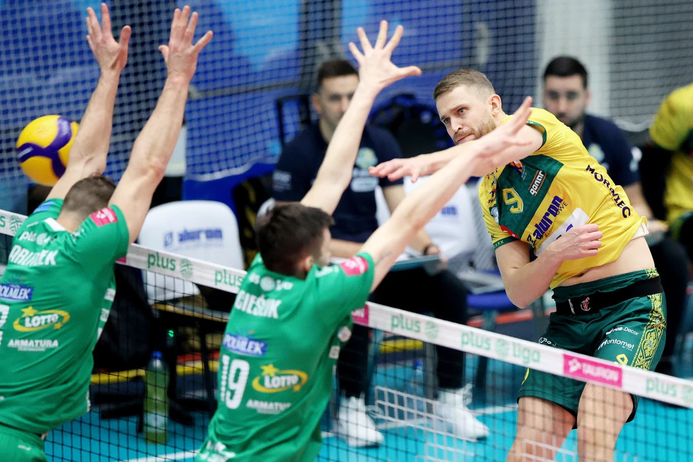 Bartłomiej Bołądź atakuje w meczu z Indykpolem AZS Olsztyn Siatkarz w żółto-zielonym stroju atakuje piłkę podczas meczu, dwaj zawodnicy drużyny w zielonych koszulkach wyskakują do bloku, widoczna siatka oraz fragmenty hali sportowej w tle.