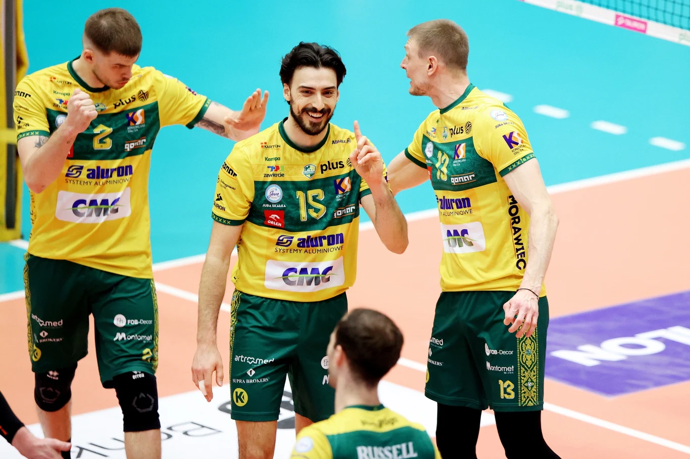 Miguel Tavares, rozgrywający Aluron CMC Warty Zawiercie Grupa siatkarzy ubranych w żółto-zielone stroje świętuje udaną akcję podczas meczu na hali sportowej, jeden z zawodników z numerem 15 w centrum cieszy się ze zdobytego punktu, otoczony drużyną.