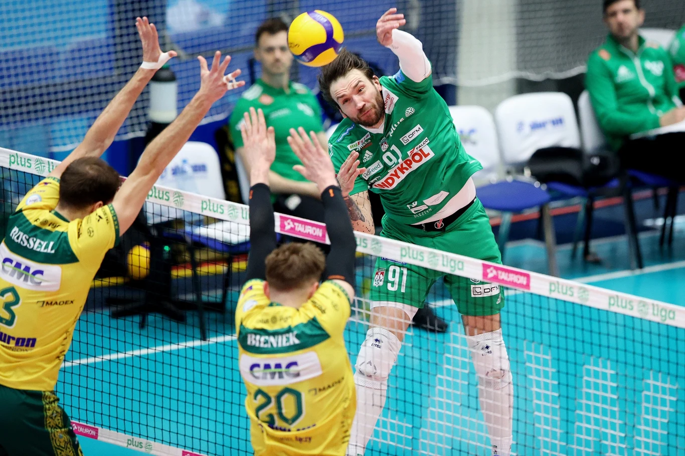 Atakuje Jan Hadrava z Indykpolu AZS Olsztyn Siatkarz w zielonym stroju wykonuje atak na piłkę, próbując przebić ją przez blok dwóch zawodników drużyny przeciwnej w żółto-zielonych strojach. W tle siatka, fragment boiska i ławka dla zawodników.