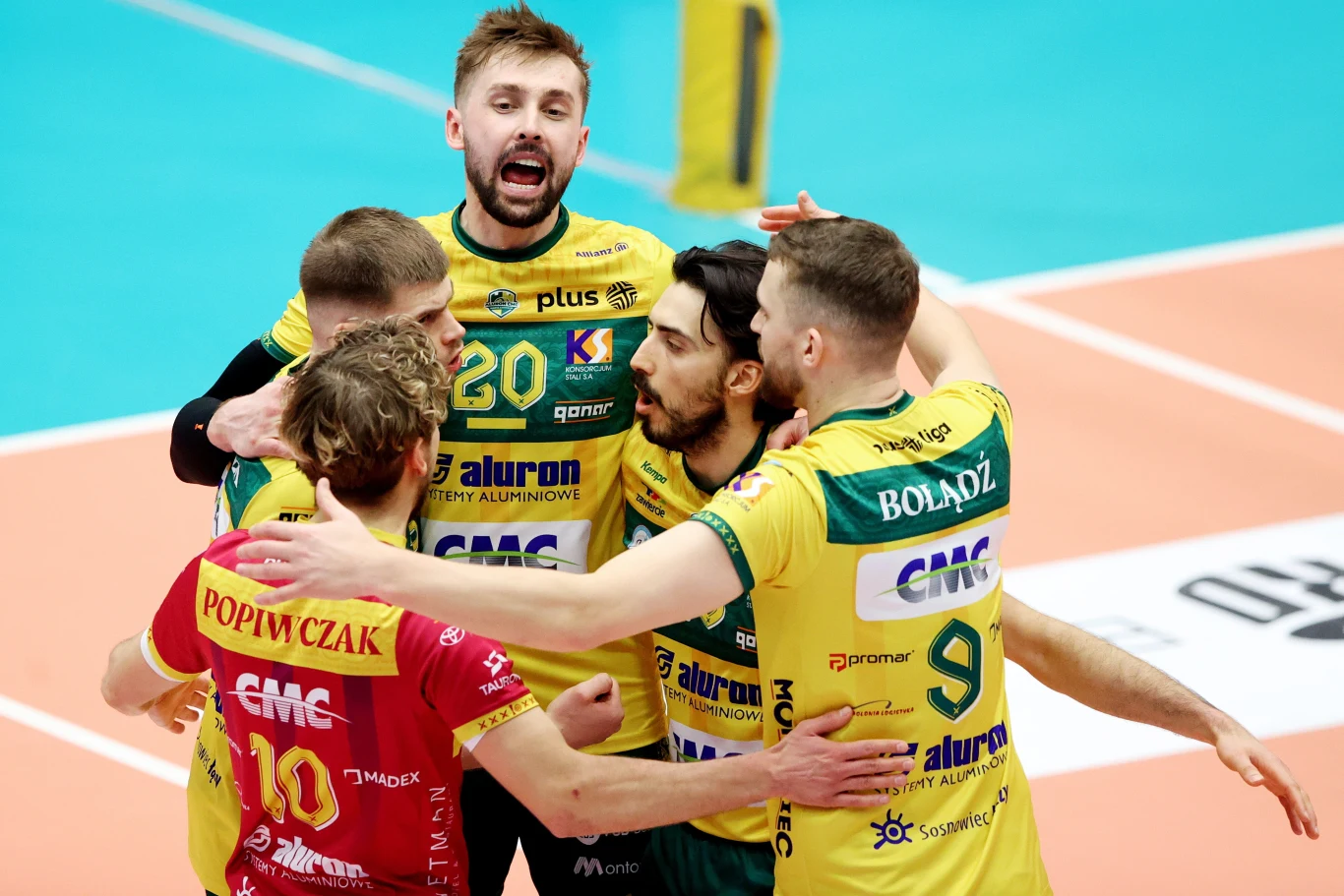 Siatkarze Aluron CMC Warty Zawiercie Grupa siatkarzy drużyny w żółto-zielonych strojach obejmuje się na boisku po zdobyciu punktu, w tle widać fragment parkietu oraz stojącego zawodnika.