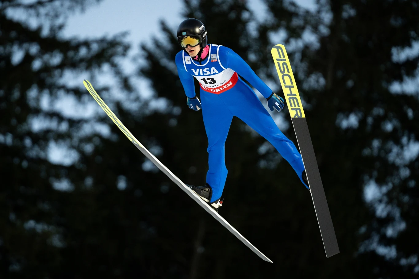 Ilja Mizernykh Skoczek narciarski ubrany w niebieski kombinezon ze startowym numerem 13 wykonuje skok, lewitując w powietrzu na tle ciemnych drzew, sprzęt sportowy widoczny na nartach.