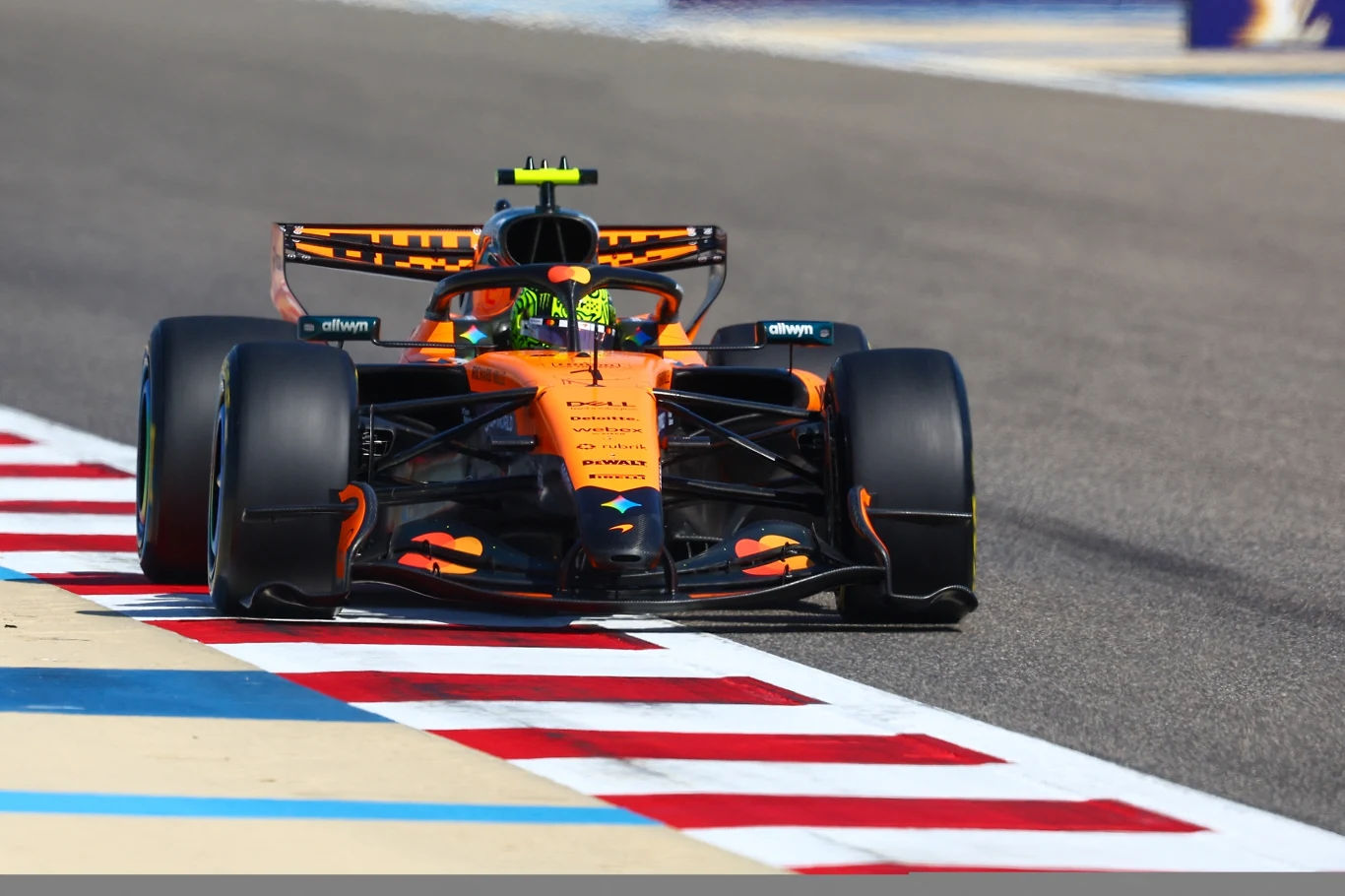 Bolid Formuły 1 w pomarańczowych barwach poruszający się po torze wyścigowym, widoczna część malowania sponsorskiego, kierowca w kasku, czerwono-białe pasy na krawędzi toru.