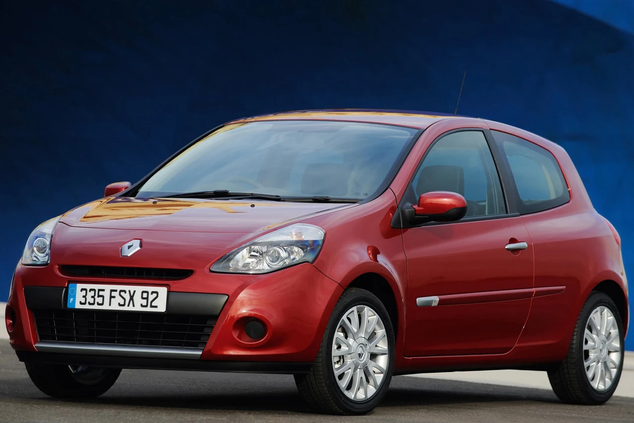 Czerwony samochód osobowy Renault Clio z francuskimi tablicami rejestracyjnymi, ustawiony pod lekkim kątem na tle niebieskiej ściany. Lśniąca karoseria i srebrne felgi podkreślają nowoczesny wygląd auta.