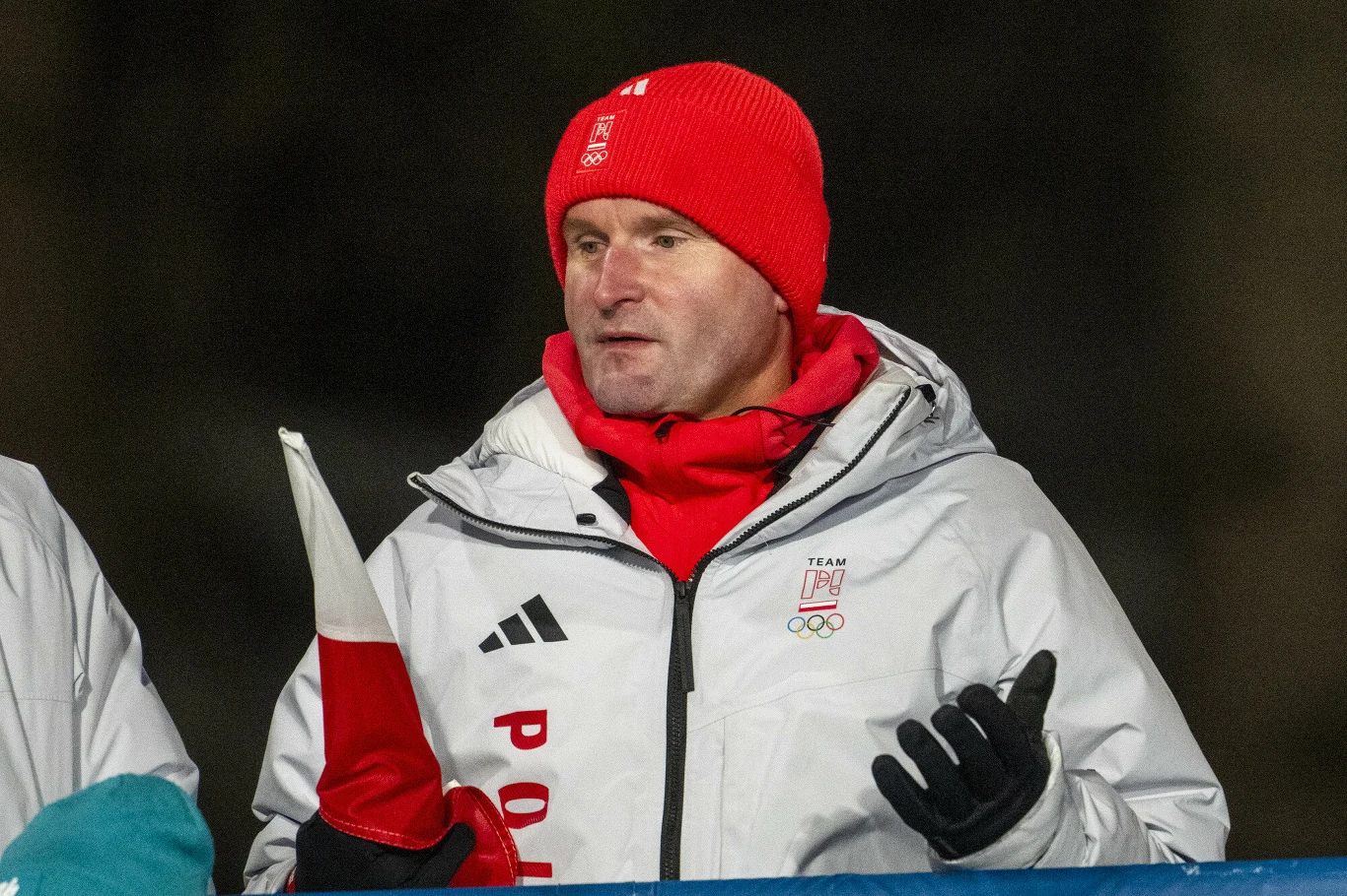 Mężczyzna w czerwonej czapce i białej kurtce z napisem Polska oraz logotypem olimpijskim trzyma polską flagę i gestykuluje podczas wydarzenia sportowego na świeżym powietrzu.