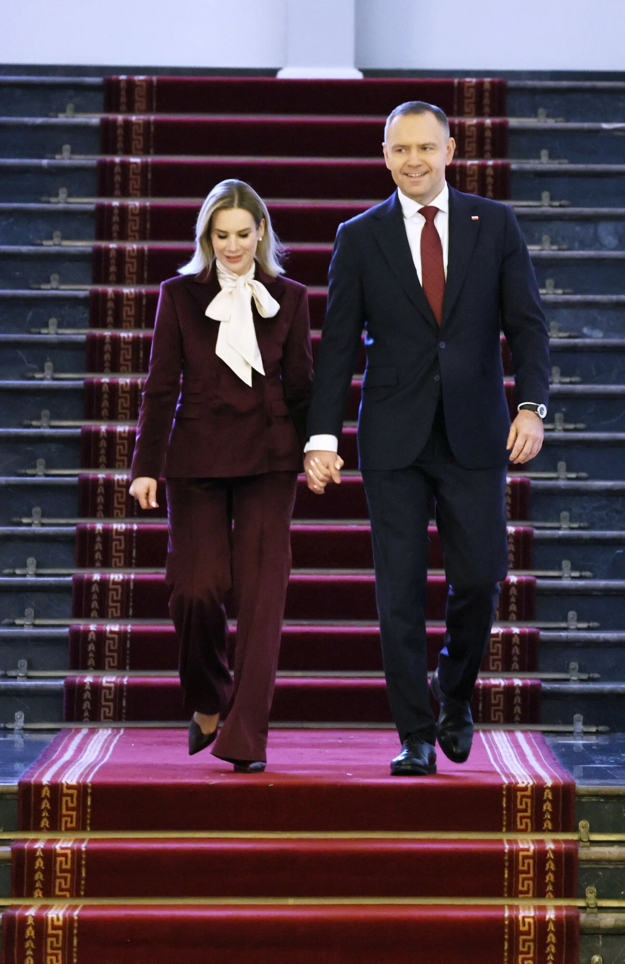 Para ubrana formalnie schodząca razem po szerokich, eleganckich schodach pokrytych czerwonym dywanem, trzymając się za ręce. Mężczyzna w ciemnym garniturze z czerwoną krawatą, kobieta w bordowym garniturze z białą kokardą pod szyją.