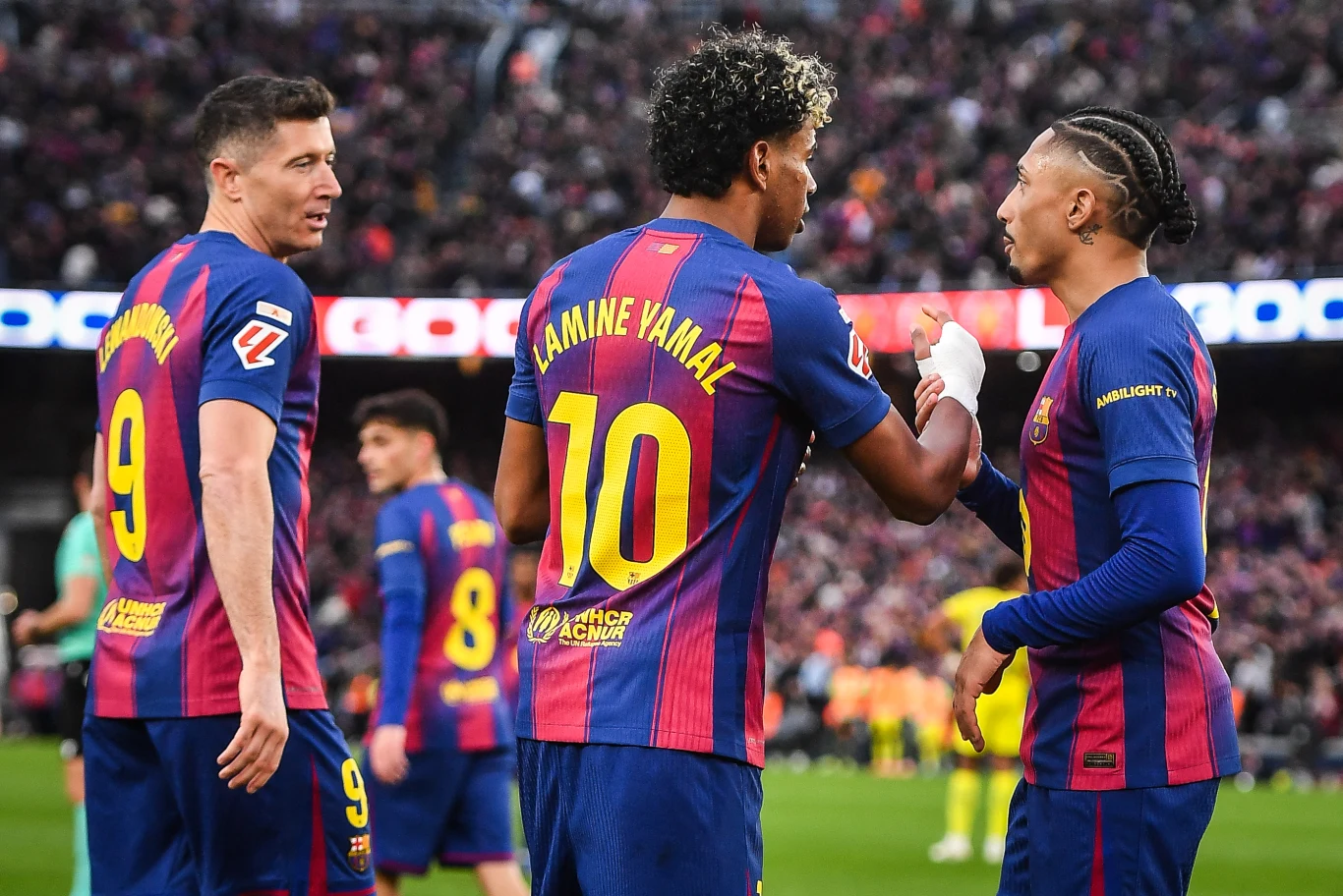 Robert Lewandowski, Lamine Yamal i Raphinha Trzech piłkarzy drużyny FC Barcelona w granatowo-bordowych strojach rozmawia na boisku podczas meczu, w tle widownia pełna kibiców i pozostali zawodnicy.