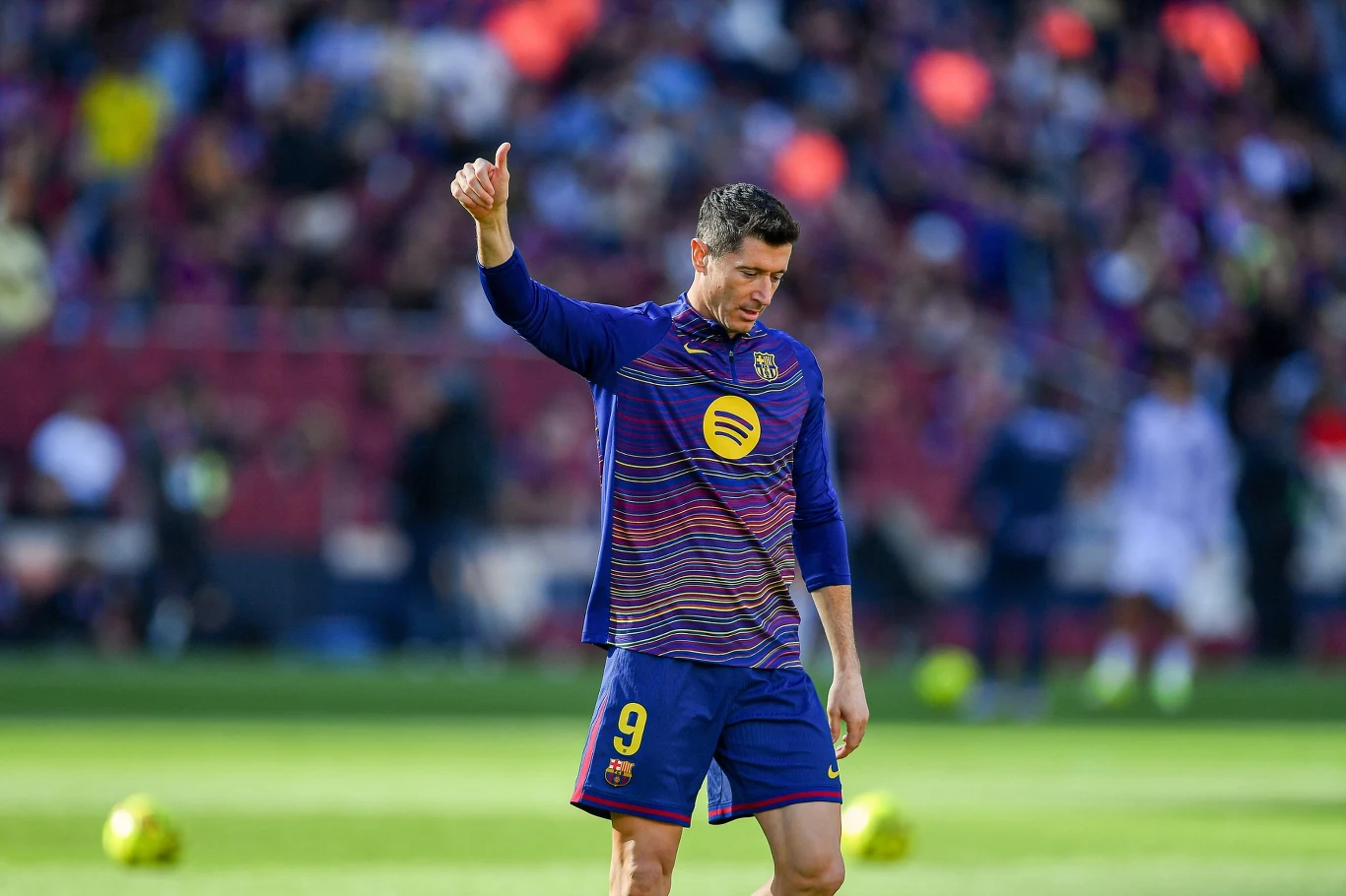 Robert Lewandowski Piłkarz ubrany w strój treningowy FC Barcelony stoi na murawie, unosi kciuk do góry, w tle widać rozmytą publiczność na stadionie oraz piłkę.
