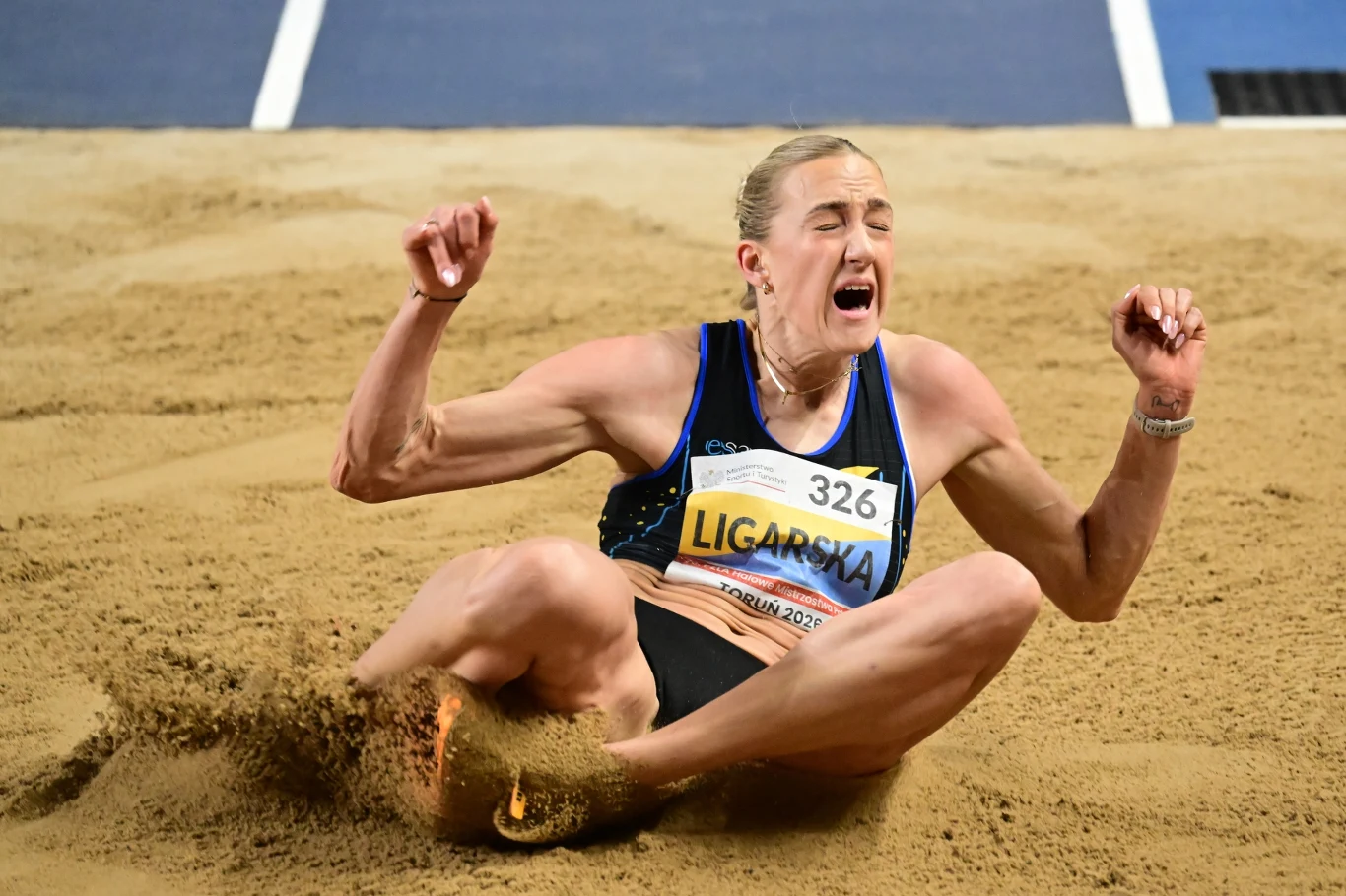 Sportowiec w trakcie lądowania podczas skoku w dal na arenie lekkoatletycznej, intensywny wyraz twarzy oraz opadający piasek podkreślają wysiłek i dynamikę momentu.