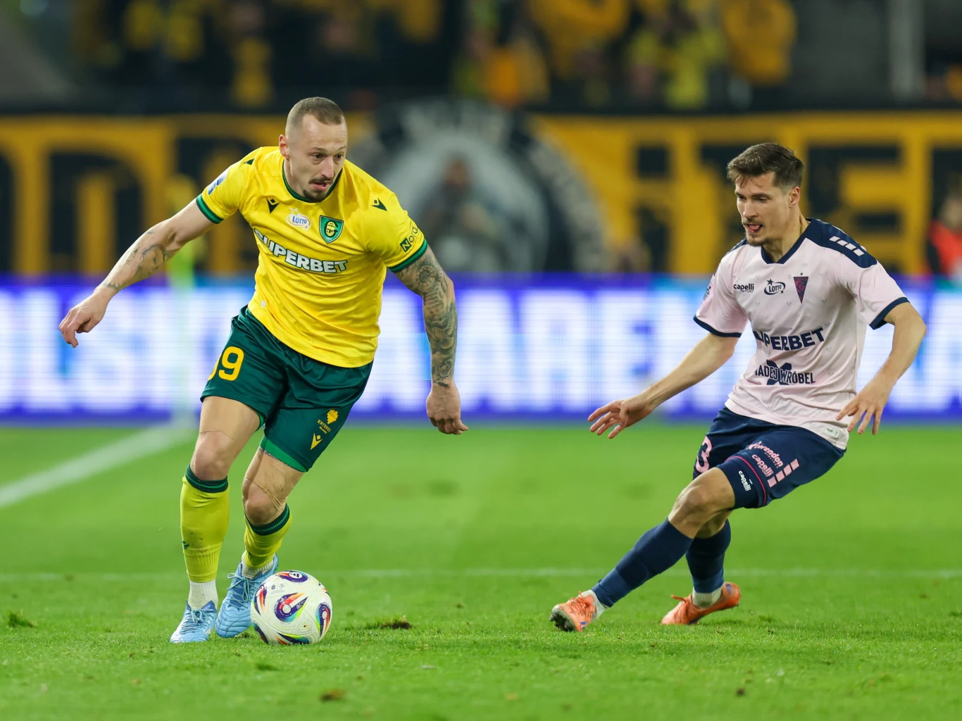 GKS-Górnik Dwóch piłkarzy walczy o piłkę na boisku, jeden ubrany w żółto-zielony strój z numerem 9, a drugi w różowo-granatowy. W tle widoczne trybuny z kibicami i banery reklamowe.
