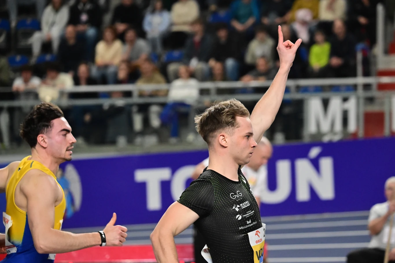 Biegacze finiszujący na bieżni podczas zawodów lekkoatletycznych w hali, jeden ze sportowców unosi rękę w geście triumfu, w tle widoczni są kibice na trybunach.