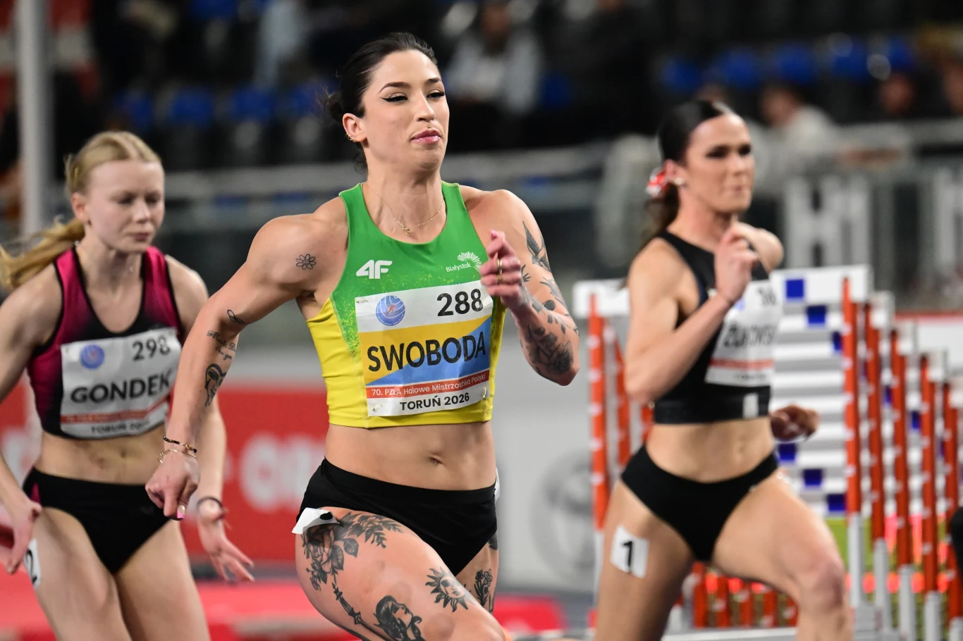 Grupa zawodniczek biegnących w wyścigu na hali, na pierwszym planie liderka w żółto-zielonym stroju z widocznymi tatuażami na udzie i ramieniu, w tle inne biegaczki oraz płotki rozstawione na torze.