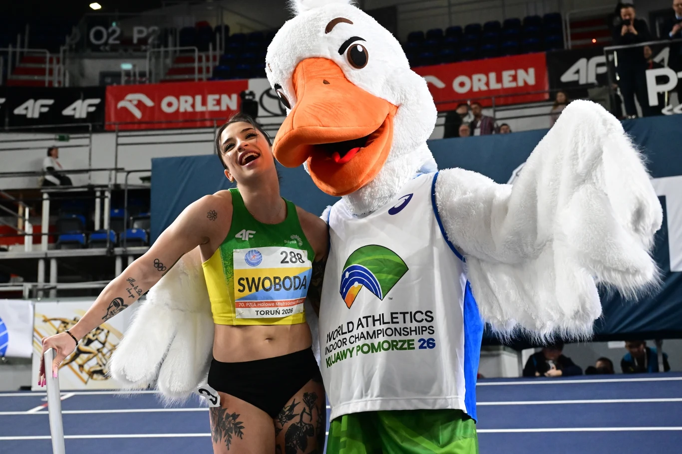 Uśmiechnięta lekkoatletka w sportowym stroju pozuje obok maskotki przypominającej dużego ptaka, oboje stoją na hali sportowej podczas wydarzenia lekkoatletycznego, w tle widać trybuny oraz banery reklamowe.