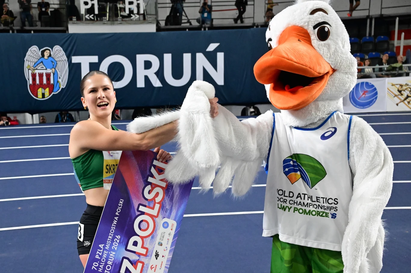 Pia Skrzyszowska Zwyciężczyni zawodów lekkoatletycznych trzyma dyplom i uśmiechając się ściska dłoń maskotki bociana w koszulce z logo mistrzostw. W tle widać trybuny oraz napis Toruń.