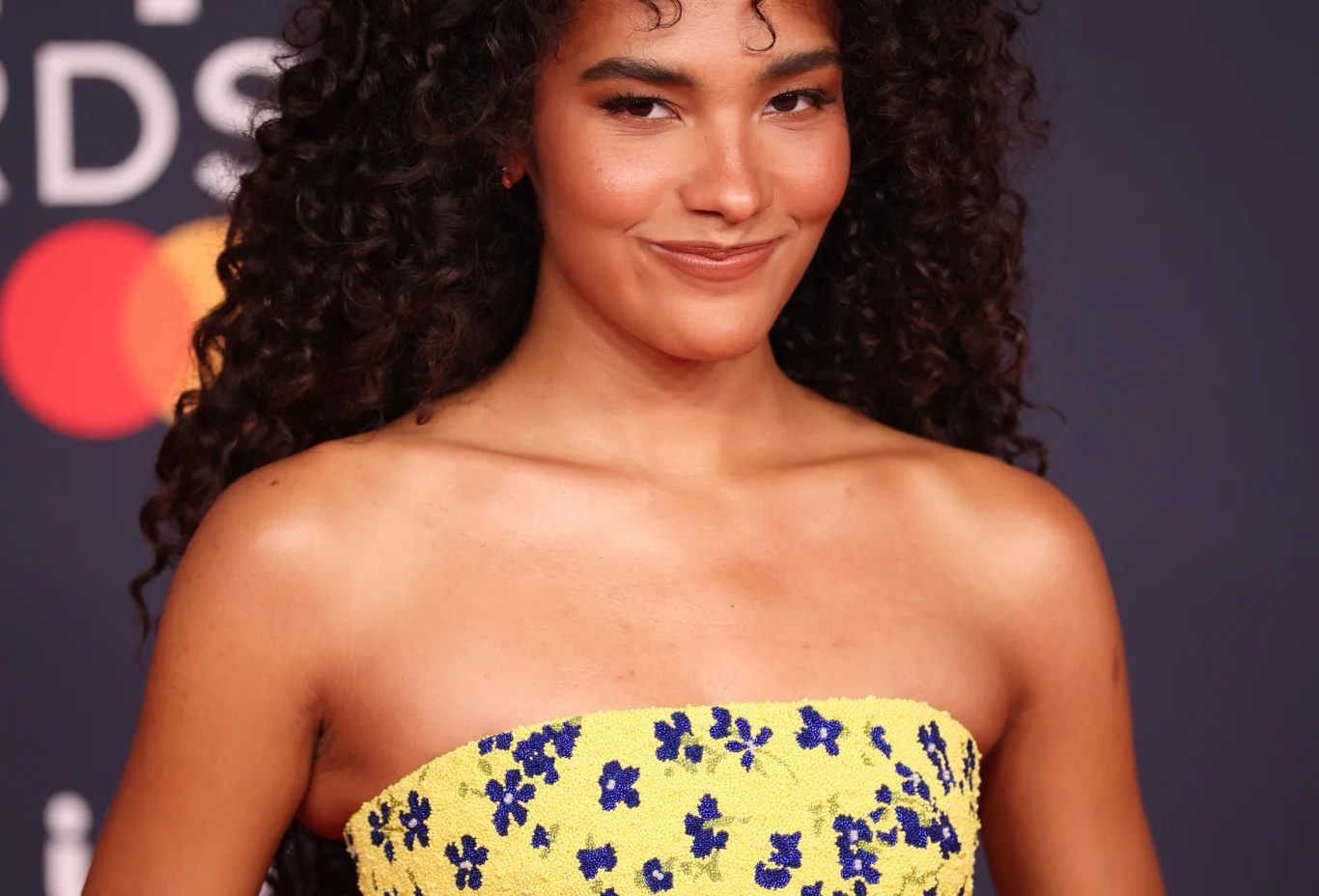 Olivia Dean zdobyła najwięcej nagród Kobieta o długich, kręconych, ciemnych włosach pozuje do zdjęcia, ubrana w żółtą sukienkę ozdobioną niebieskimi kwiatami, na tle z logotypem Mastercard.