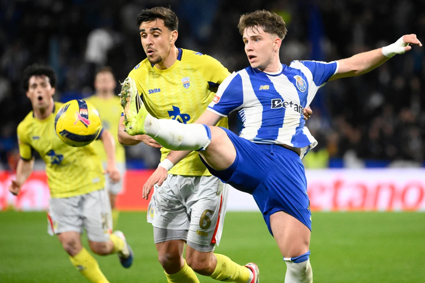 Dwóch piłkarzy walczy o piłkę w dynamicznej akcji na boisku podczas meczu, jeden z zawodników unosi nogę, aby przejąć piłkę, a w tle widać innych graczy i rozmytą publiczność.