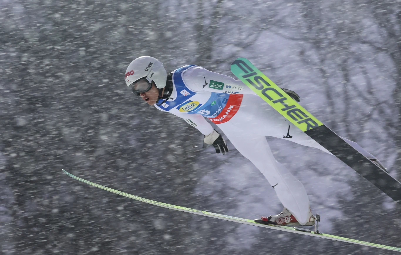 Skoczek narciarski w trakcie lotu przez śnieżną zamieć, ubrany w biały kombinezon i kask, z charakterystycznymi nartami przedstawiającymi markę Fischer, dynamiczna sylwetka na tle rozmytego, zimowego krajobrazu.