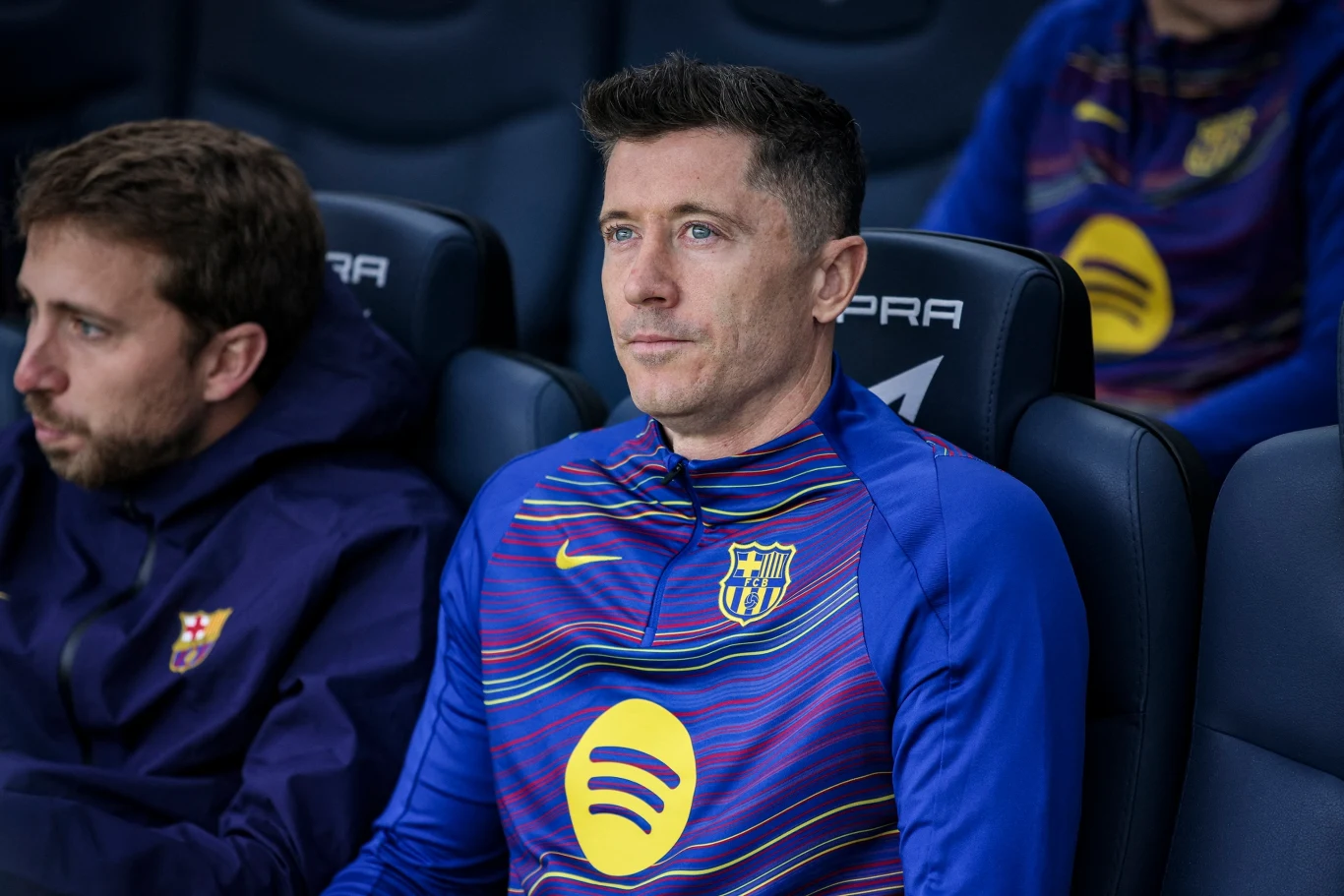 Robert Lewandowski Piłkarz ubrany w klubowy dres FC Barcelony z logo sponsora na klatce piersiowej siedzi na ławce rezerwowych, patrzy przed siebie, obok niego inny członek drużyny.