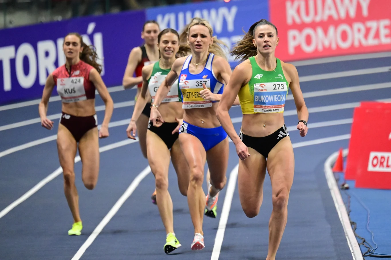 Finisz finału 400 metrów kobiet. Natalia Bukowiecka ze złotem Grupa zawodniczek biegnie po bieżni podczas zawodów halowej lekkoatletyki, na czele stawki znajduje się kobieta w zielonym stroju z numerem 273, w tle widoczne są trybuny i banery reklamowe.