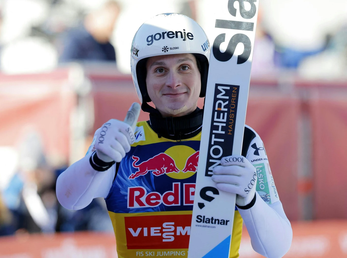 Domen Prevc Skoczek narciarski w kombinezonie startowym i kasku, stojący z nartą w dłoni, uśmiechnięty pokazuje kciuki w górę. W tle widoczny fragment trybun i elementy infrastruktury zawodów.
