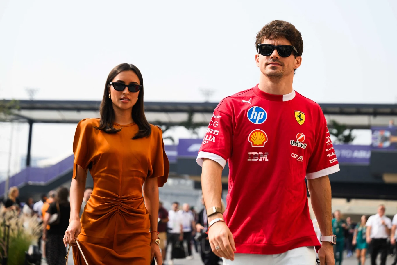 Alexandra Saint Mleux i Charles Leclerc Mężczyzna w koszulce Ferrari i okularach przeciwsłonecznych idzie obok kobiety w eleganckiej, pomarańczowej sukience, oboje są otoczeni przez innych ludzi na tle obiektów sportowych.
