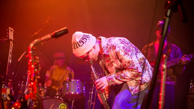 Grupa muzyków grających na scenie podczas koncertu, z głównym akcentem na osobę w kwiecistej koszuli, białej czapce i okularach grającą na saksofonie. W tle perkusista oraz gitarzysta podświetleni kolorowymi światłami.