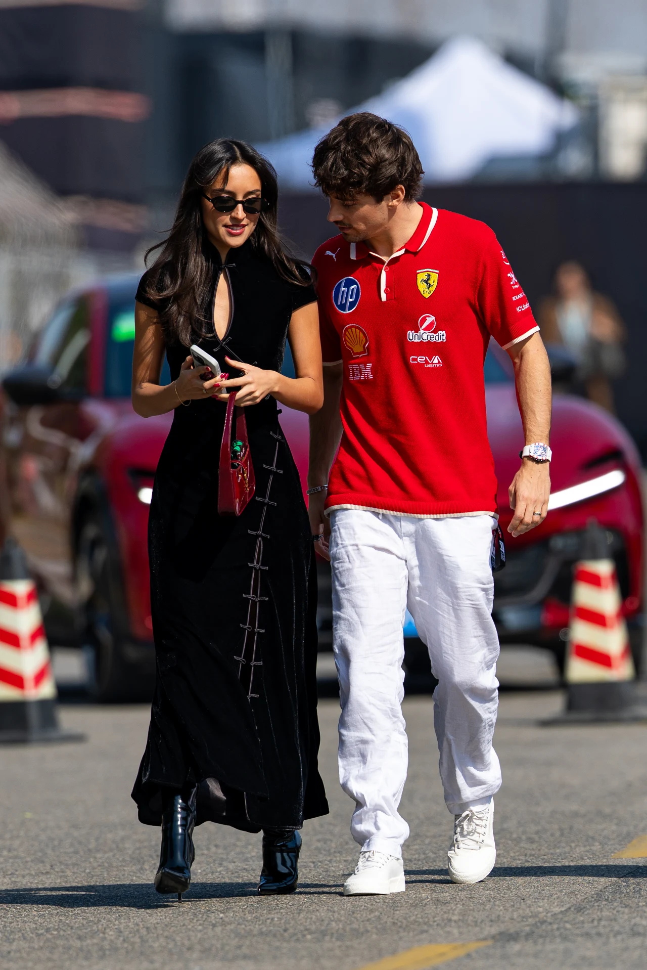 Alexandra Saint Mleux i Charles Leclerc Dwie osoby spacerują obok siebie, kobieta w długiej czarnej sukience i mężczyzna w czerwonej koszulce Ferrari oraz białych spodniach, w tle stoją czerwone samochody oraz widoczne są prowizoryczne pachołki drogowe.