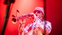 Mężczyzna w ciemnych okularach, kolorowej koszuli i czapce gra na saksofonie na tle czerwonego światła scenicznego.