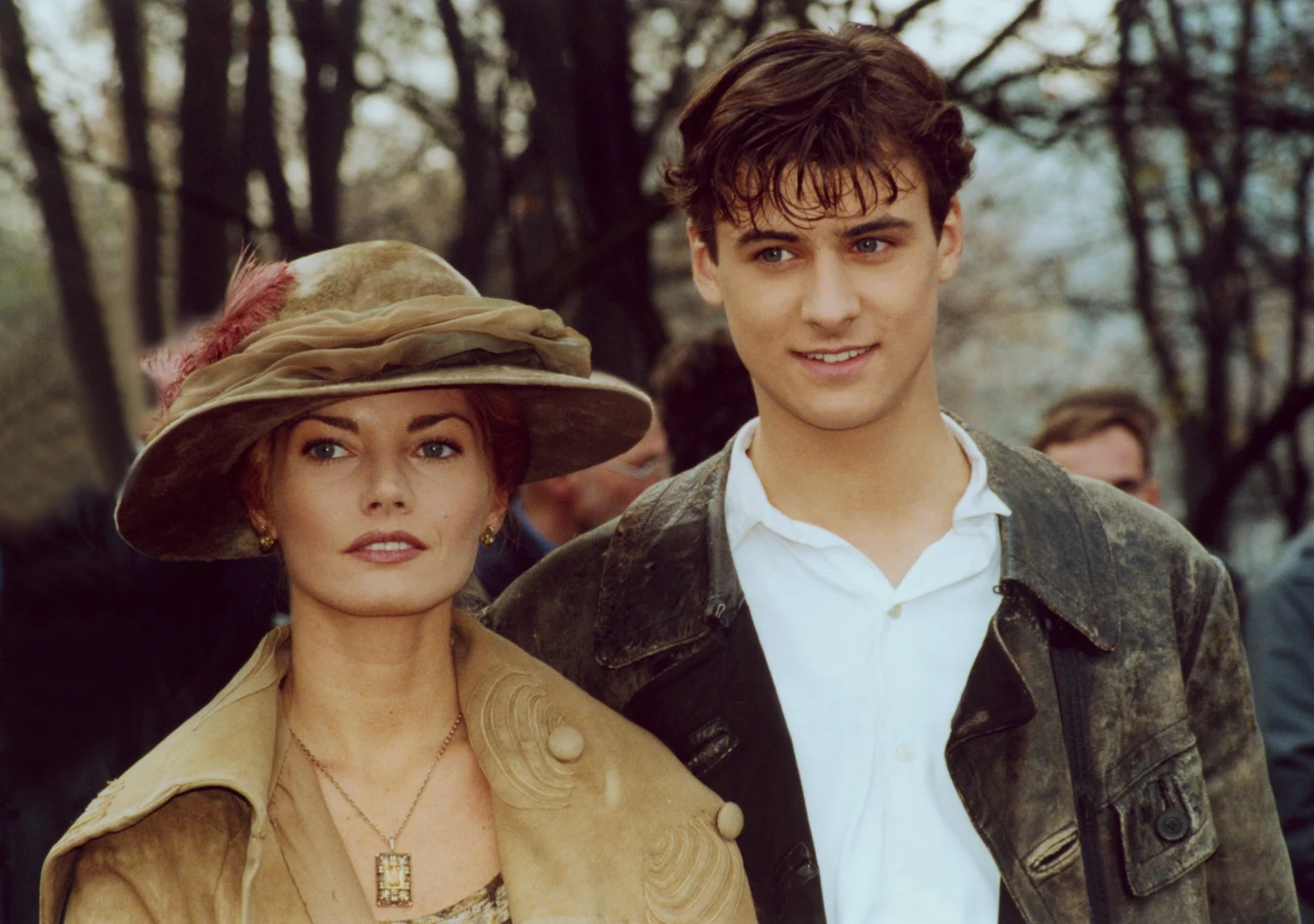 Małgorzata Lewińska i Mateusz Damięcki w filmie "Przedwiośnie" Kobieta w eleganckim kapeluszu i zdobionym płaszczu stoi obok młodego mężczyzny ubranego w białą koszulę i ciemną kurtkę, w tle widać rozmyte sylwetki ludzi i drzewa.
