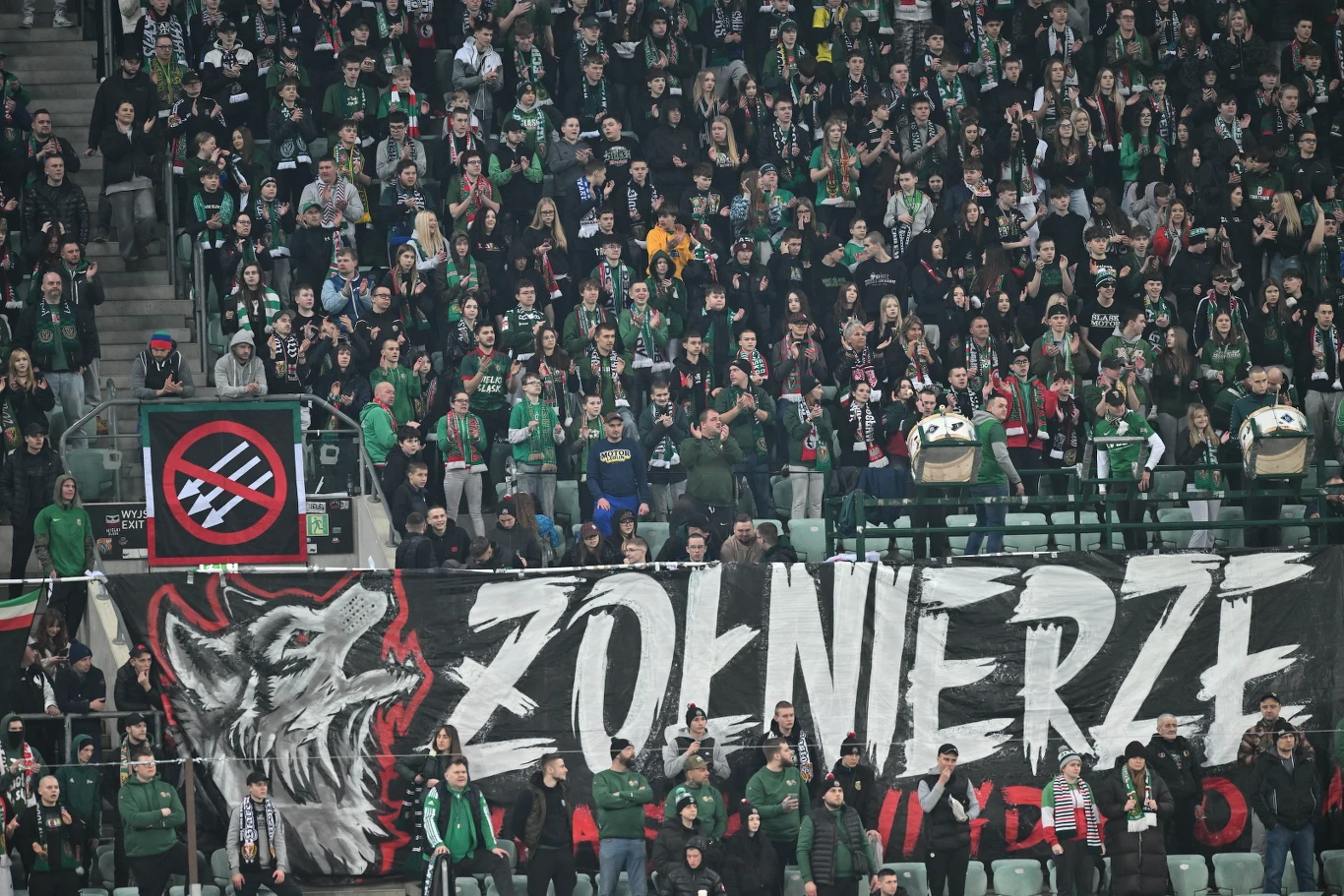 Tragedia na stadionie we Wrocławiu Tłum kibiców na stadionie z dużym transparentem z napisem 'Żołnierze' oraz wizerunkiem wilka, z dominującymi barwami zieleni i czerni, liczne flagi oraz gesty uniesionych rąk.