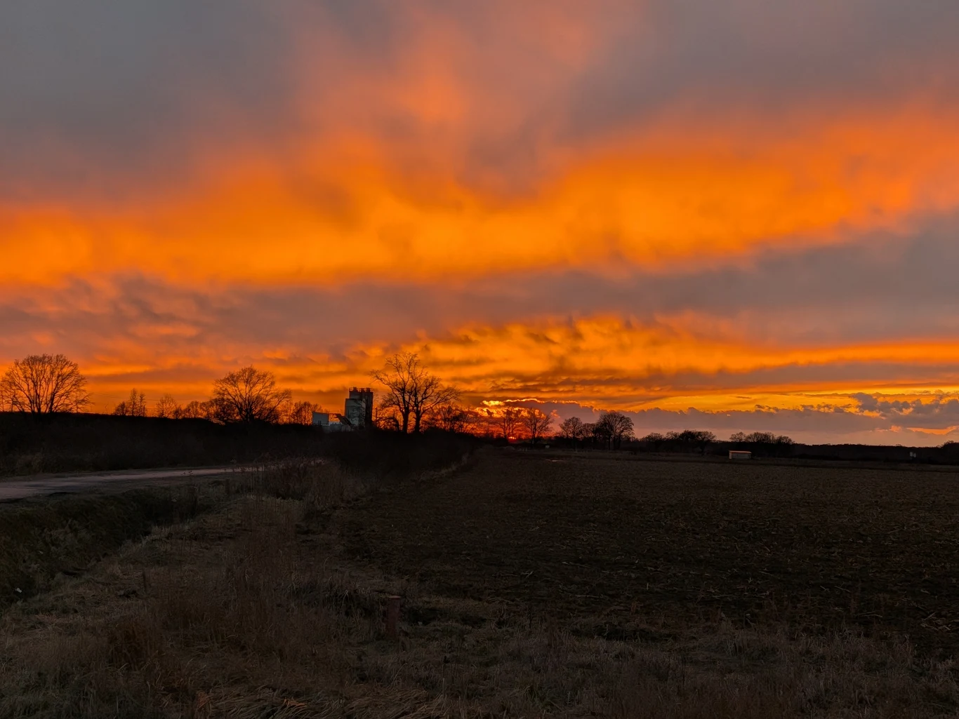 Pola uprawne na pierwszym planie, w oddali rzucające się w oczy pomarańczowe i żółte barwy nieba podczas zachodu słońca, sylwetki drzew i niewielki budynek na horyzoncie.