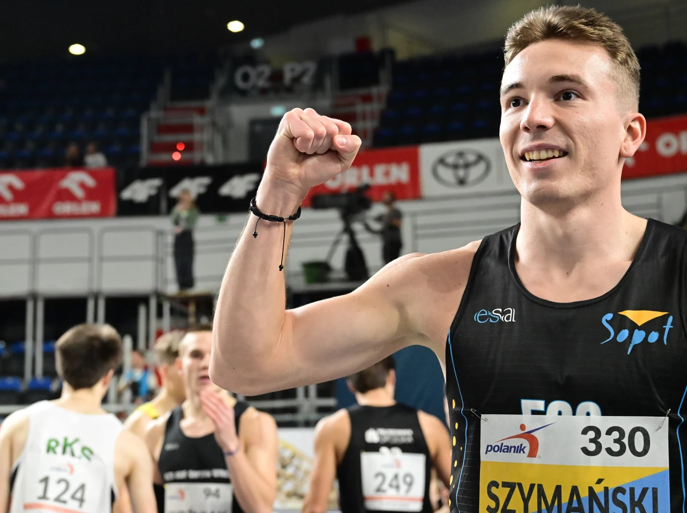 Młody sportowiec z numerem startowym 330 i nazwiskiem Szymański na piersi świętuje swój sukces, unosząc zaciśniętą pięść w geście triumfu. W tle widoczni są inni uczestnicy biegu oraz trybuny areny lekkoatletycznej.